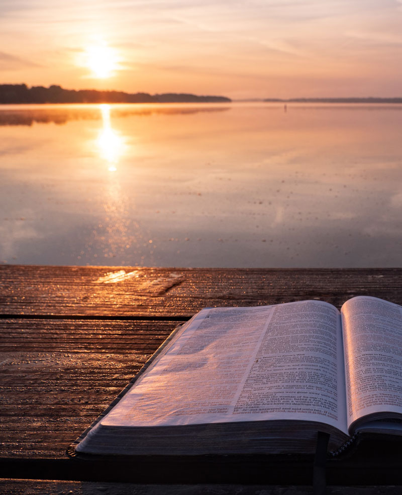 An open Bible on the shore of the lake with the sun setting in beautiful hues of pinks and oranges